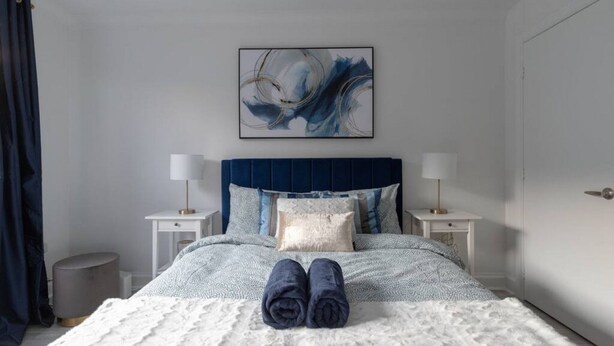 A neatly made bed with a blue headboard, two bedside tables with lamps, a large abstract painting above the bed, and a pair of folded towels on the bed.