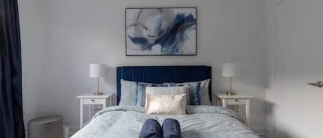 A neatly made bed with a blue headboard, two bedside tables with lamps, a large abstract painting above the bed, and a pair of folded towels on the bed.