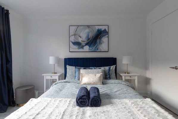 A neatly made bed with a blue headboard, two bedside tables with lamps, a large abstract painting above the bed, and a pair of folded towels on the bed.