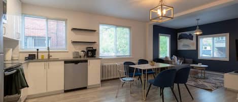 A modern kitchen with a dining area, large windows, and a ceiling light fixture.