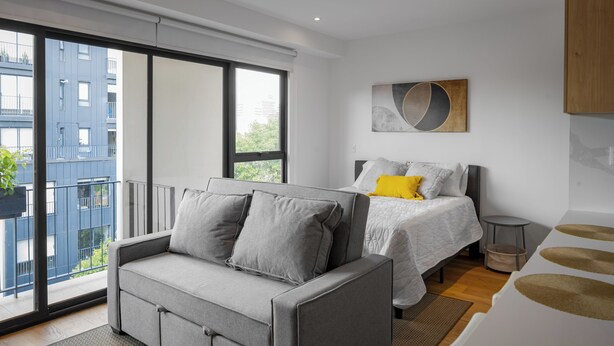 A modern bedroom with a grey sofa, a bed with white bedding, and a view of a building through large windows. - Reforma Elegance · A Pasos del Monumento & Ángel (Ciudad de México)
