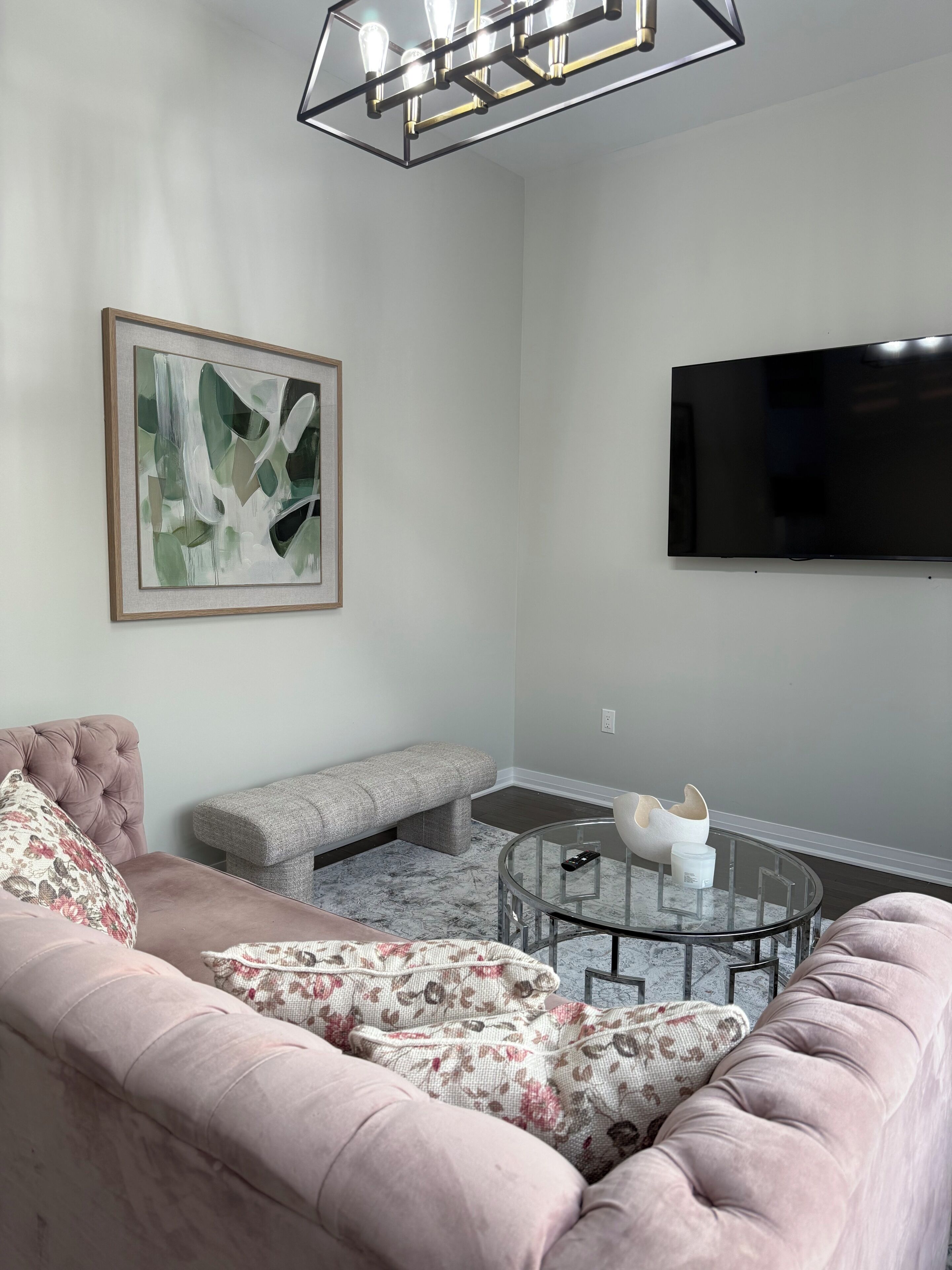 A living room with a sofa, bench, round glass coffee table, abstract painting, and a flat-screen TV.