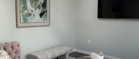 A living room with a sofa, bench, round glass coffee table, abstract painting, and a flat-screen TV.