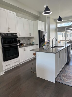 A modern kitchen with white cabinetry, stainless steel appliances, and a large island with a sink. - Luxury Waterfront Retreat | Walk to Beach | Sleeps 18 (Clarington)