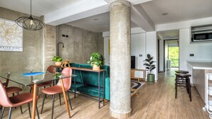 Um interior moderno com área de jantar, mesa de vidro, cadeiras vermelhas e sofá verde. É visível um pilar grande e um lustre.