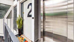 Entrada de um edifício com uma porta marcada com o número 2, um vaso de planta, um portão de metal e um tapete de entrada com a palavra 'WELCOME'.