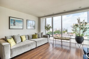 Una sala de estar moderna con un sofá gris, dos cuadros enmarcados, un ventanal con vista a la ciudad y una planta en maceta.