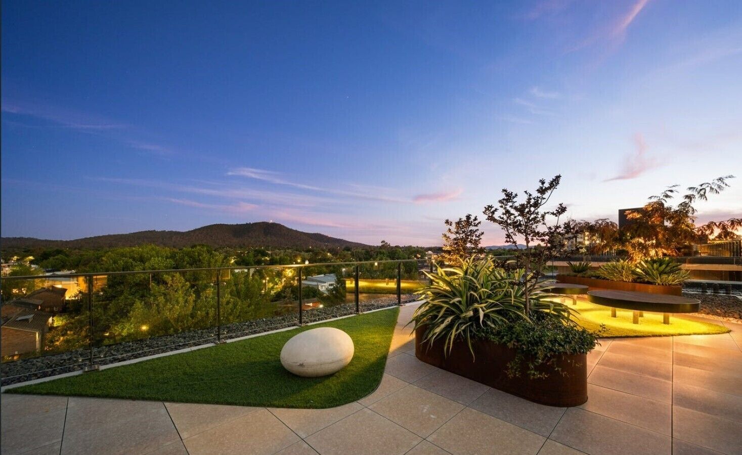 A rooftop terrace with a view of a cityscape and hills, featuring a seating area with planters and a spherical bench.