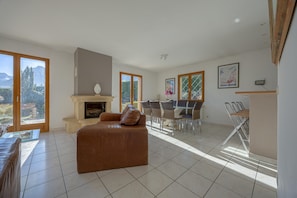 A spacious living room with a fireplace, a dining area with a glass table, and large windows offering a view of the outdoors. - Beautiful Family Home with a Pool, Nestled Between the Lake and the Mountains (Lathuile)