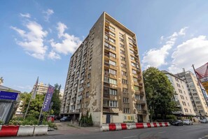 Un edifici residencial de diverses plantes amb balcons, una zona d'aparcament i un cel clar.