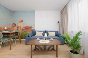 Un salon avec un canapé bleu, une table basse en bois, une table à manger avec des chaises et un grand tableau accroché au mur.