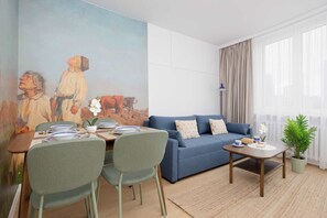 A living room with a blue sofa, a dining table with chairs, a painting on the wall, and a window with curtains. - Stylish Apartment with Loggia | 2 Bedrooms | Workspace (Warszawa)