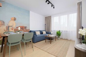 A modern living room with a blue sofa, dining table with chairs, a painting on the wall, and a large window with curtains. - Stylish Apartment with Loggia | 2 Bedrooms | Workspace (Warszawa)