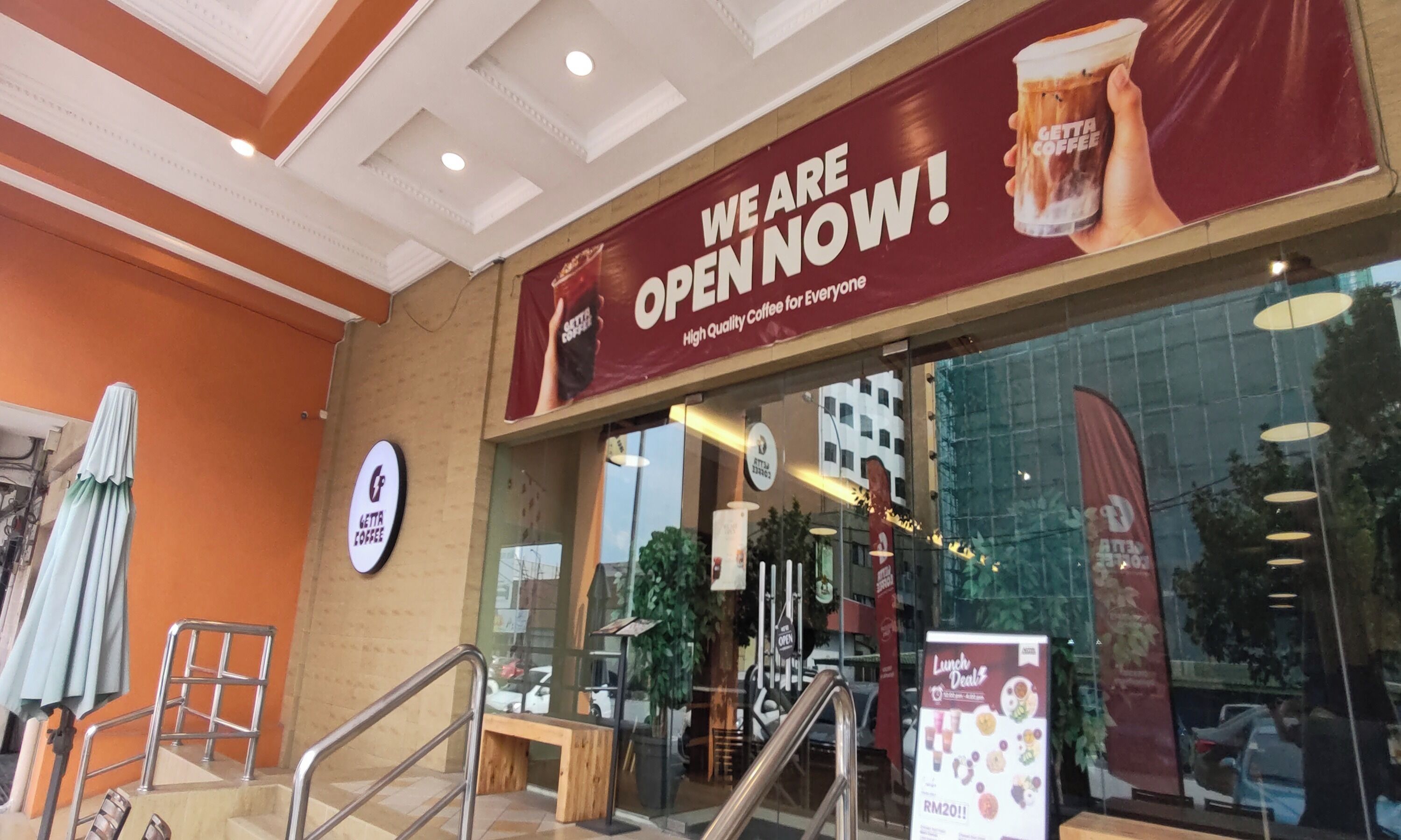 A coffee shop entrance with a prominent 'WE ARE OPEN NOW!' sign, a glass storefront, and a 'Lunch Deals' poster.