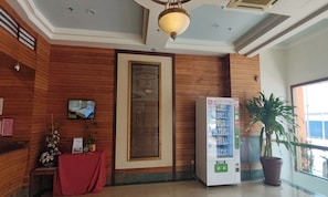 Een receptiegedeelte met een houten wand, een televisie, een automaat, een potplant en een rode tafel met een certificaat.