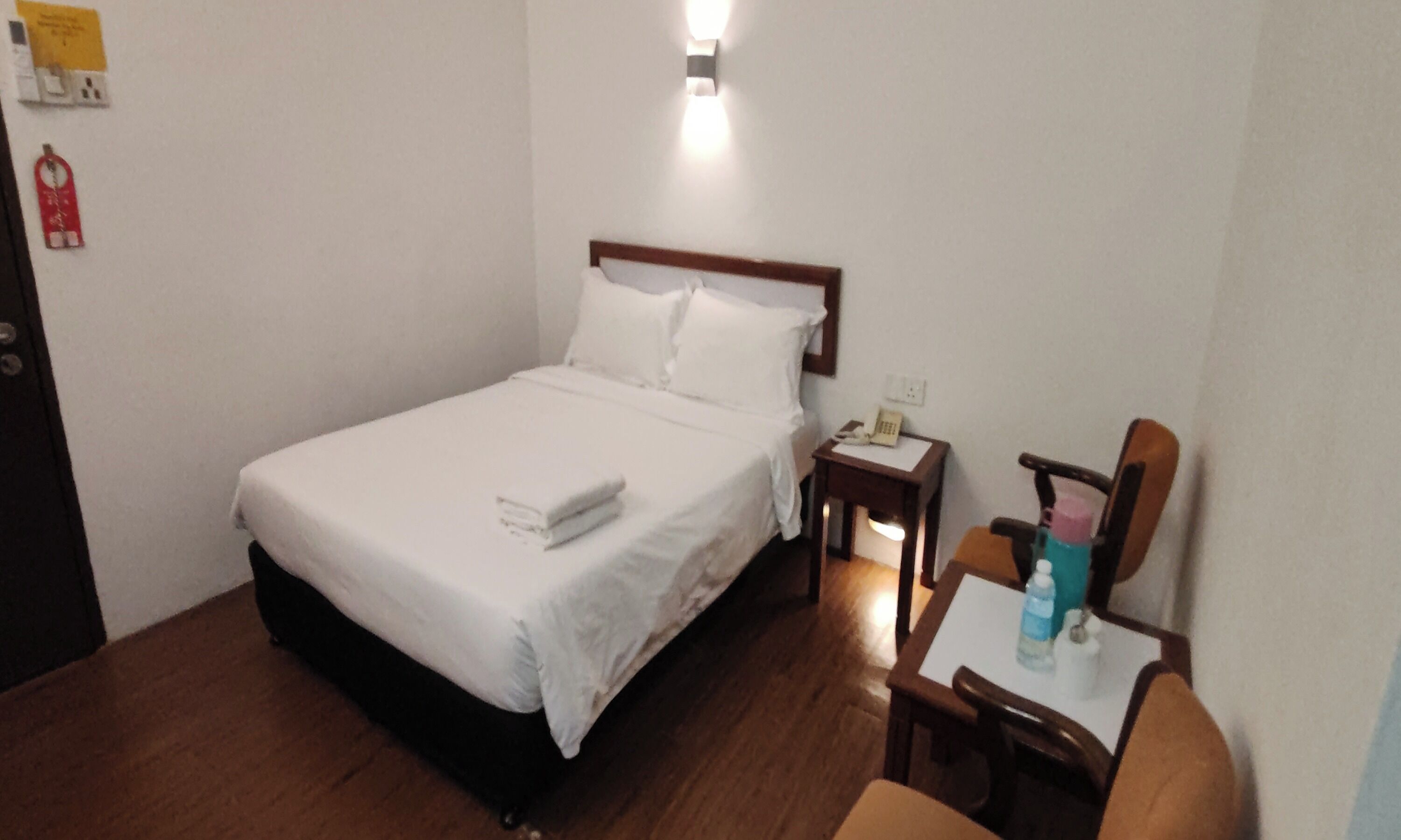 A hotel room with a bed, a nightstand, a chair, and a table with a bottle and a tissue box.