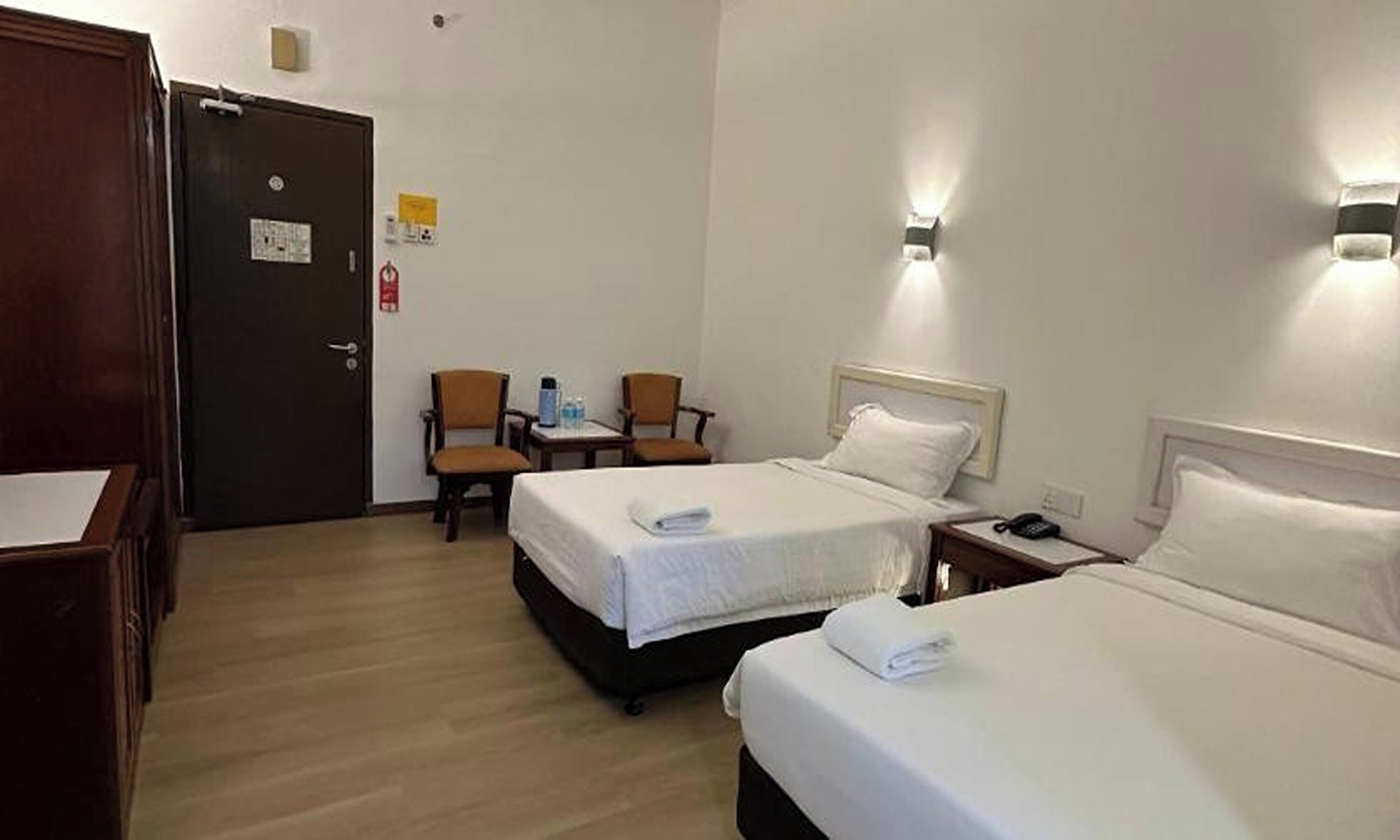Habitación de hotel con dos camas, un escritorio, dos sillas, una puerta y una lámpara fijada a la pared.
