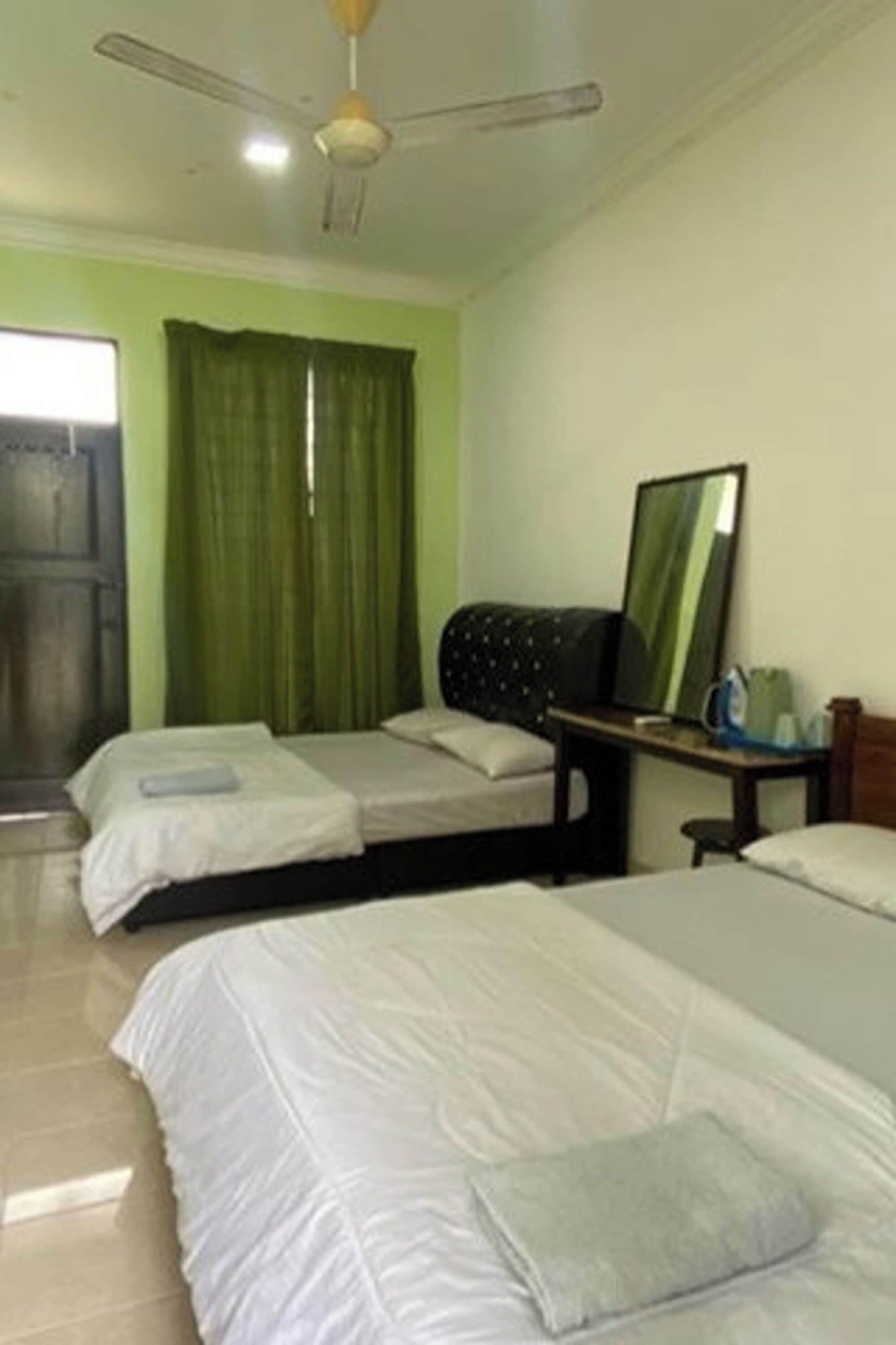 A room with two beds, a ceiling fan, a window with curtains, and a table with a mirror and a kettle.