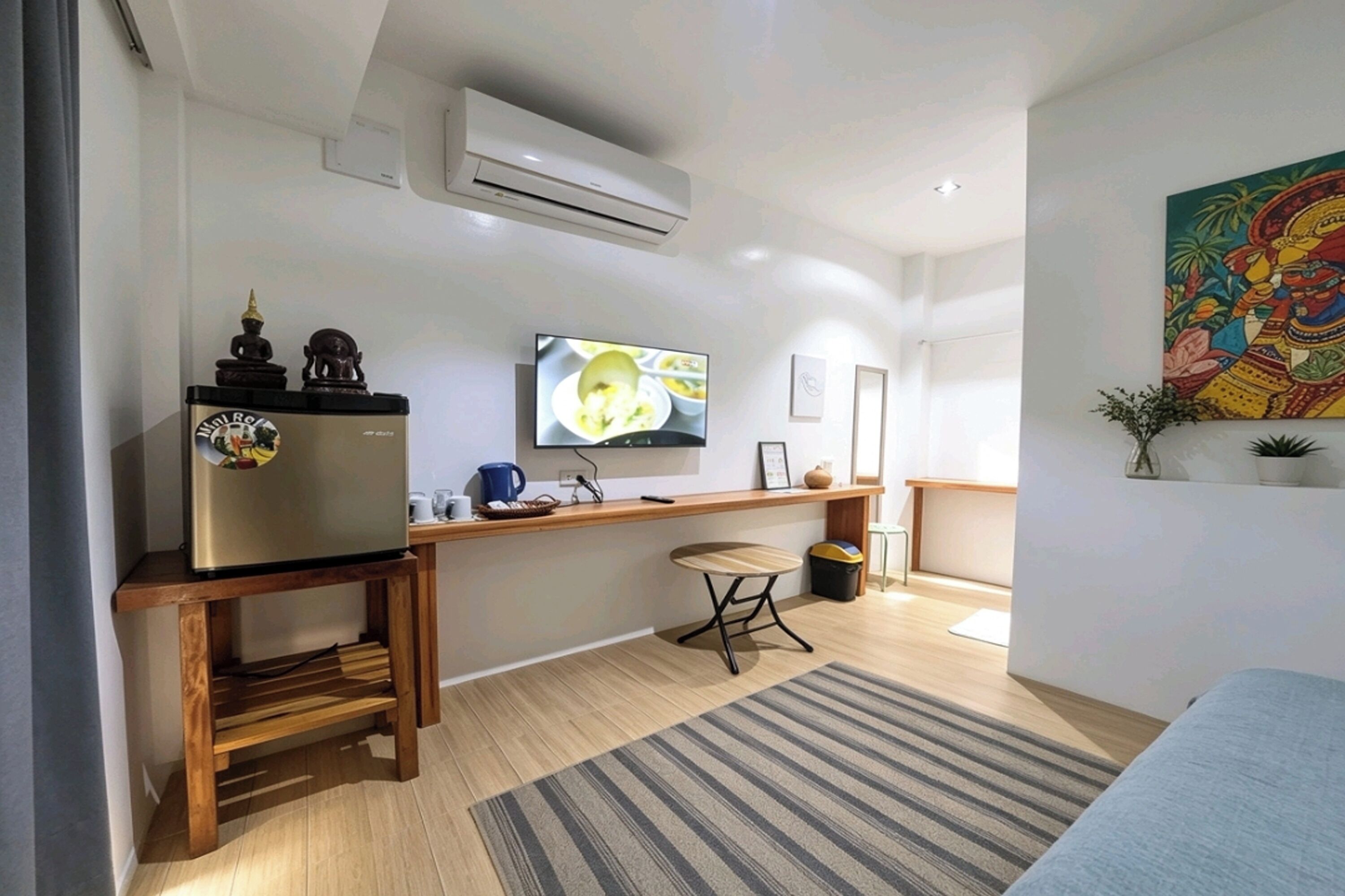 A compact living space with a kitchenette, a flat-screen TV mounted on the wall, a small wooden table, a single chair, and a colorful artwork on the wall.