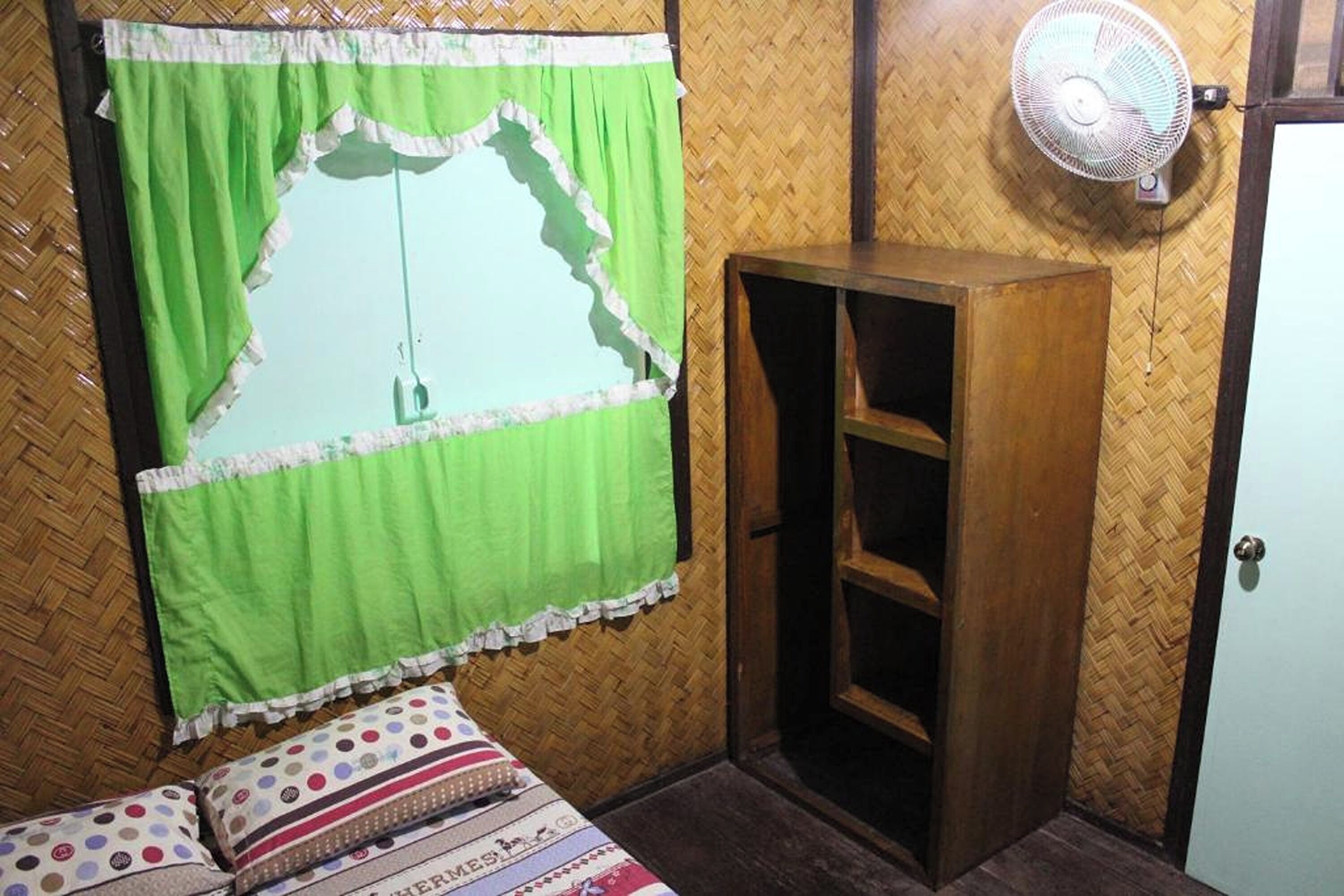 A room with a bed, a wooden wardrobe, a fan, and a window with a green curtain.