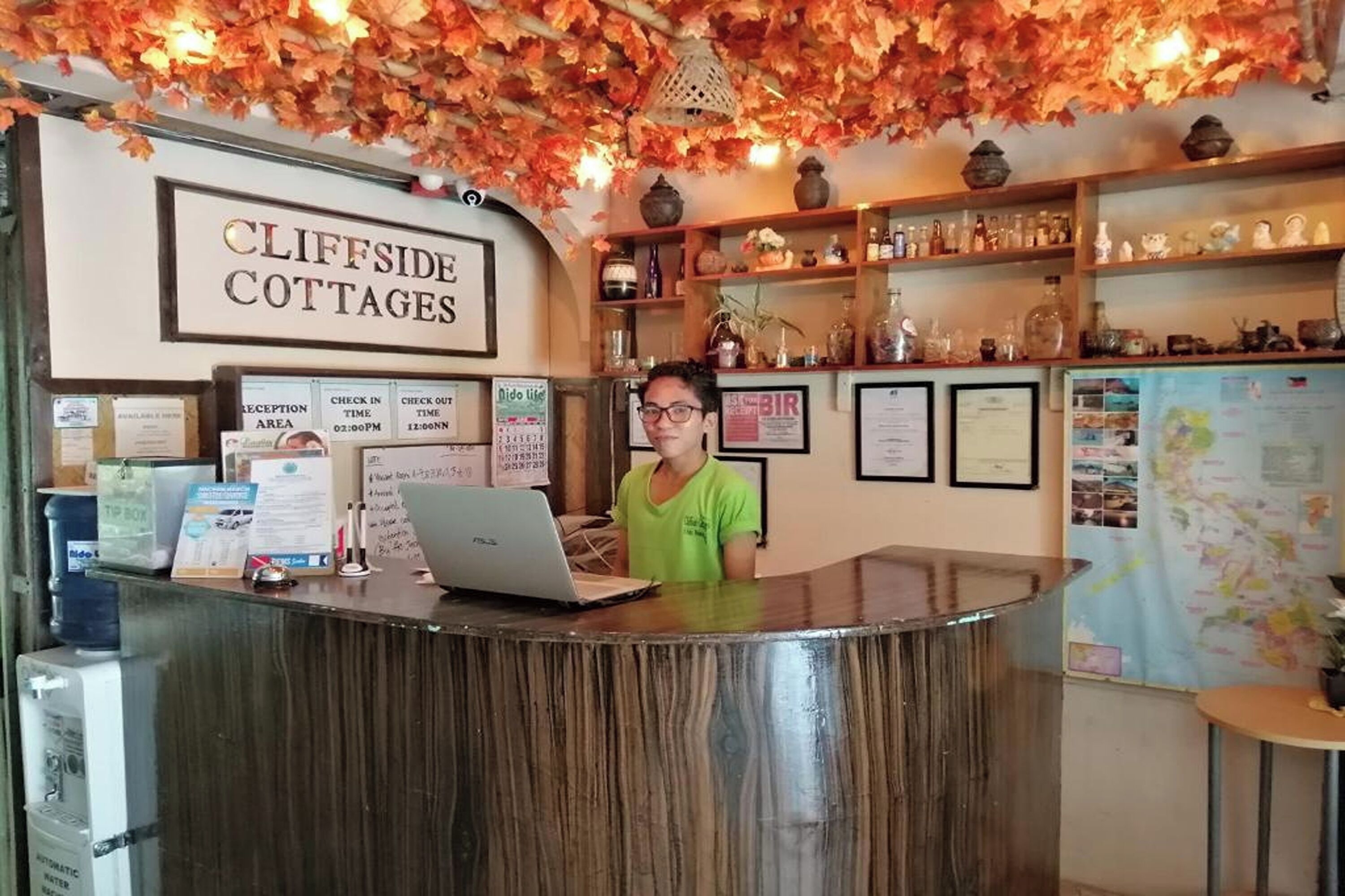 A reception desk at Cliffside Cottages with a laptop, a water dispenser, and various signs including check-in and check-out times.