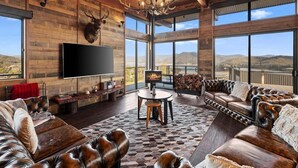 A spacious living room with a large flat-screen TV, leather sofas, a chandelier, and a fireplace. - Ridge Way Views - Breathtaking Lake Eildon Views (Mountain Bay)