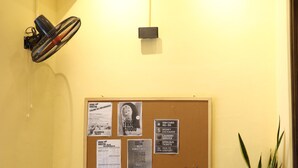 A room with a seating area, a bulletin board with various notices, a potted plant, and a wall-mounted fan. - Rebel Rooster Hostel (Hanoi)