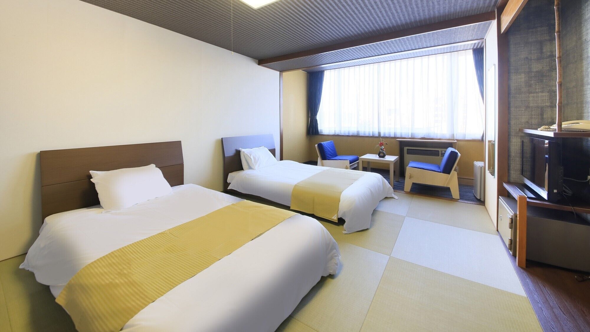 A hotel room with two beds, a small table, a chair, a TV, and a window with curtains.
