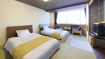Habitación de hotel con dos camas, una mesita pequeña, una silla, televisor y ventana con cortinas.