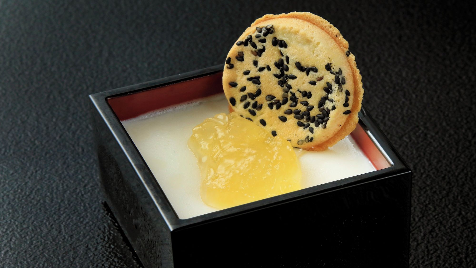 A square black box containing a cookie with black sesame seeds and a piece of yellow butter.