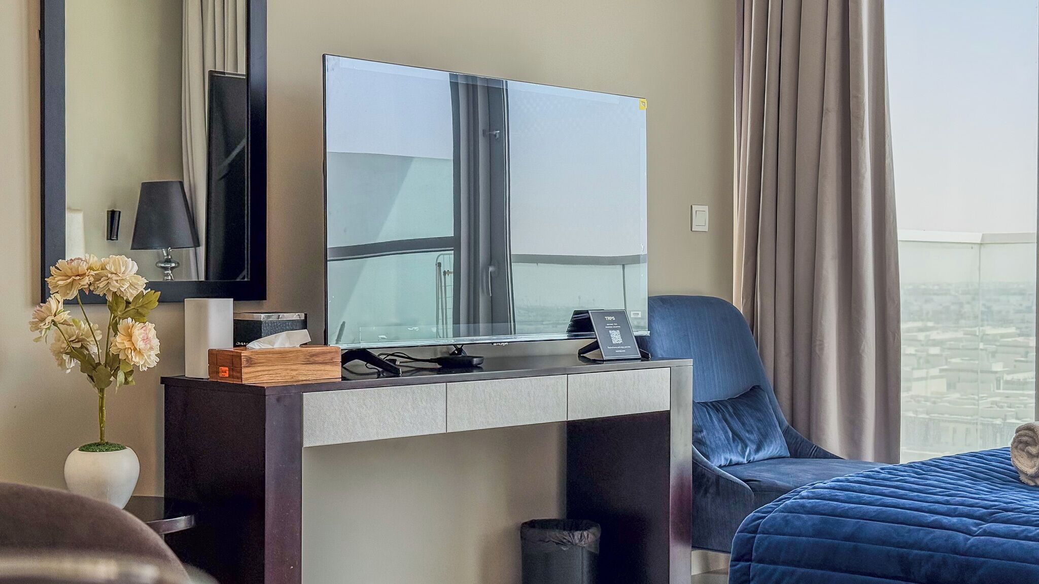 A hotel room with a bed, a desk with a flat-screen TV, a chair, a vase with flowers, and a view of the city.