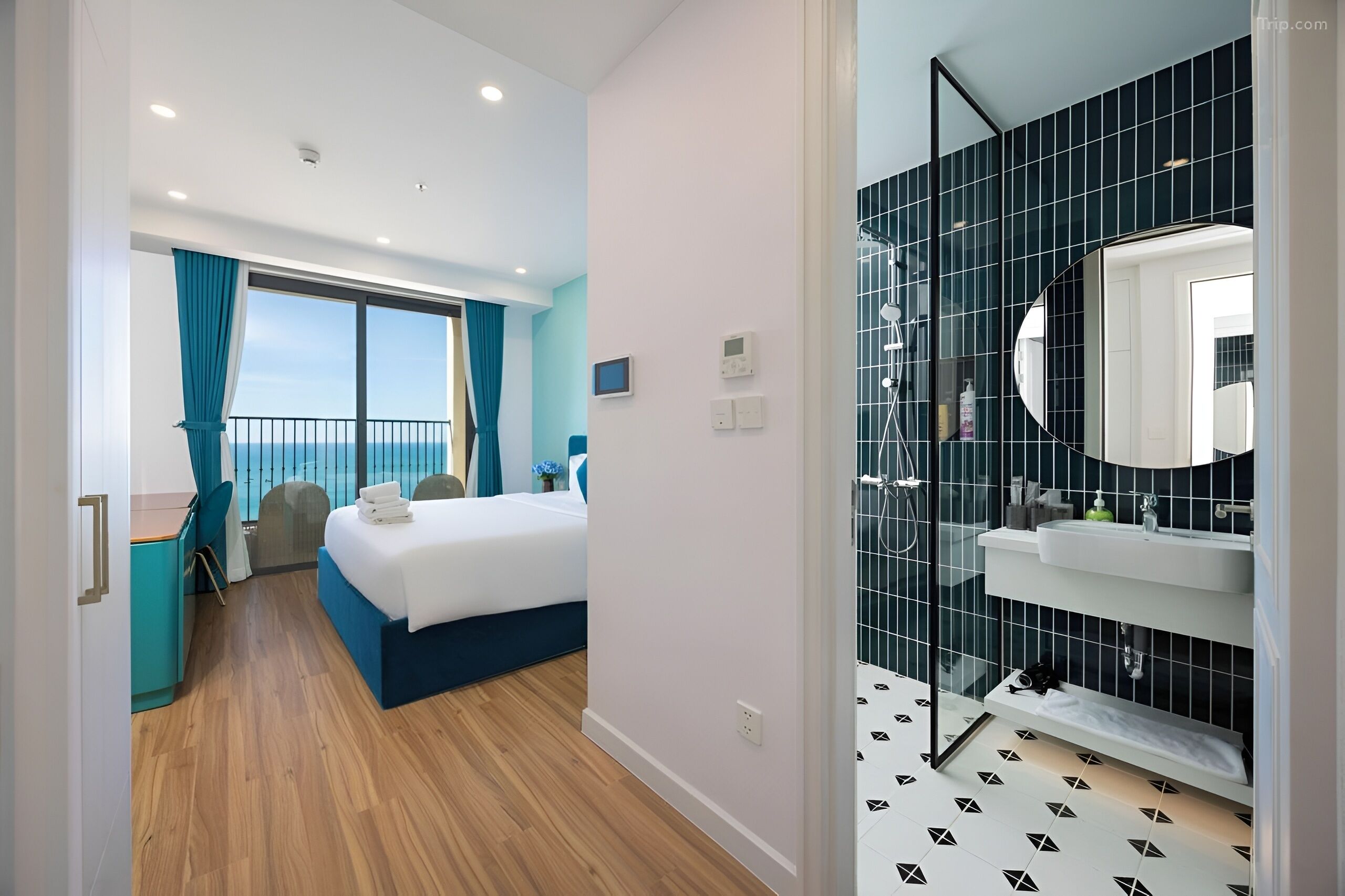 Une chambre d’hôtel moderne dotée d’un balcon, d’un lit, d’un bureau et d’une salle de bain avec un grand miroir et des murs recouverts de carreaux noirs.