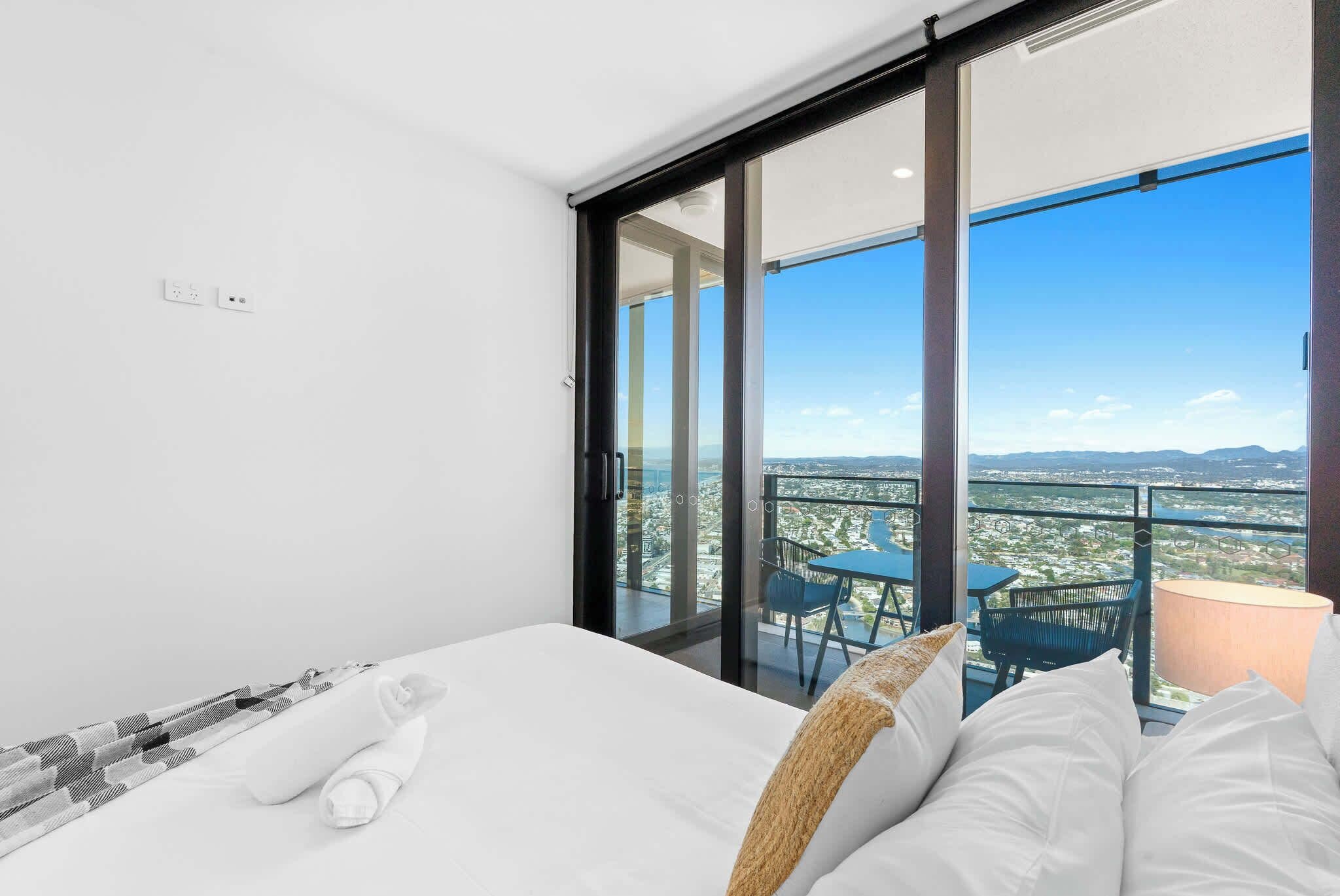 A modern hotel room with a large bed, a view of a cityscape through floor-to-ceiling windows, and a balcony with outdoor furniture.
