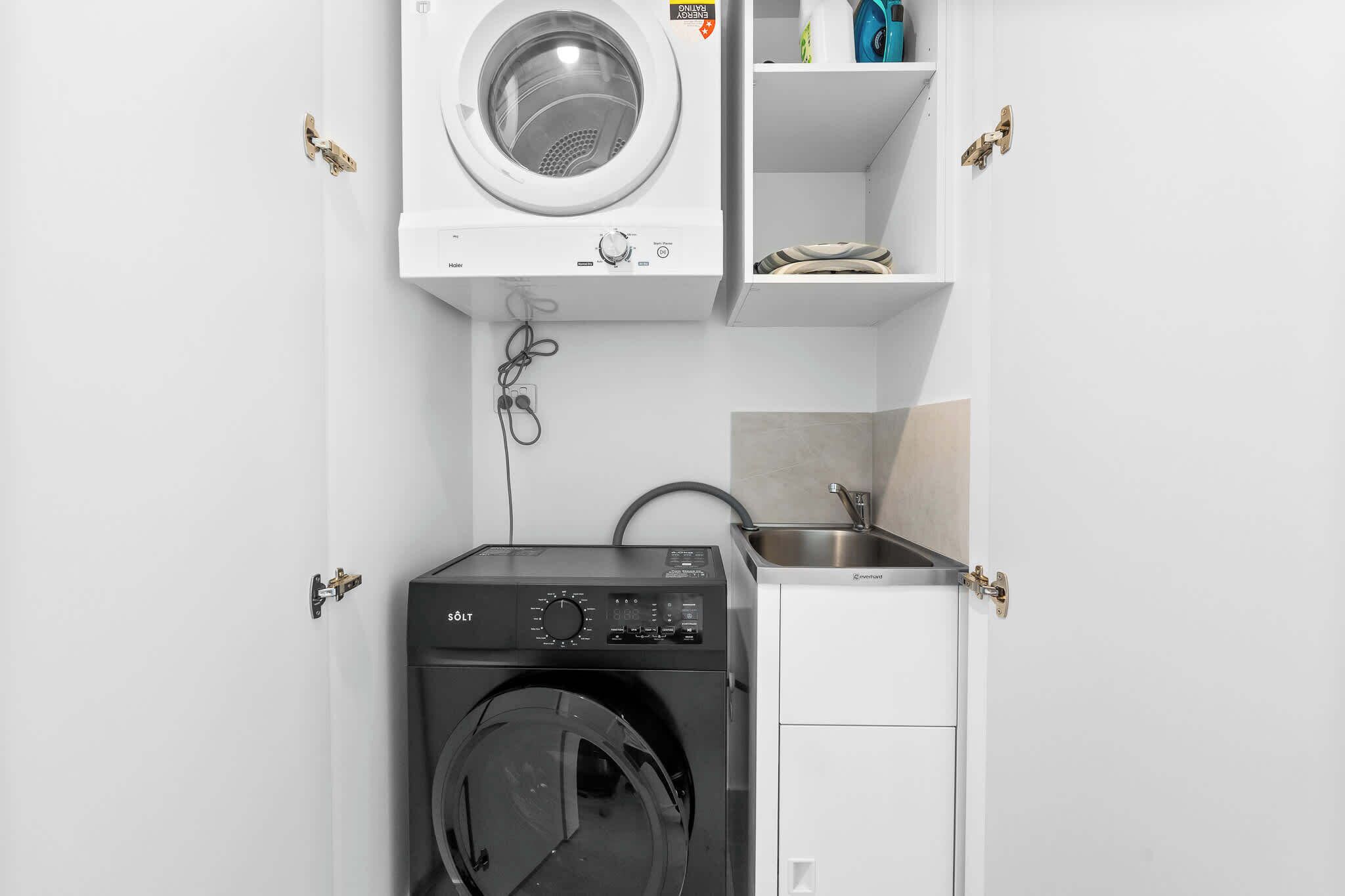 Un espace buanderie compact comprenant un lave-linge et un sèche-linge à chargement frontal, un évier et des étagères de rangement.