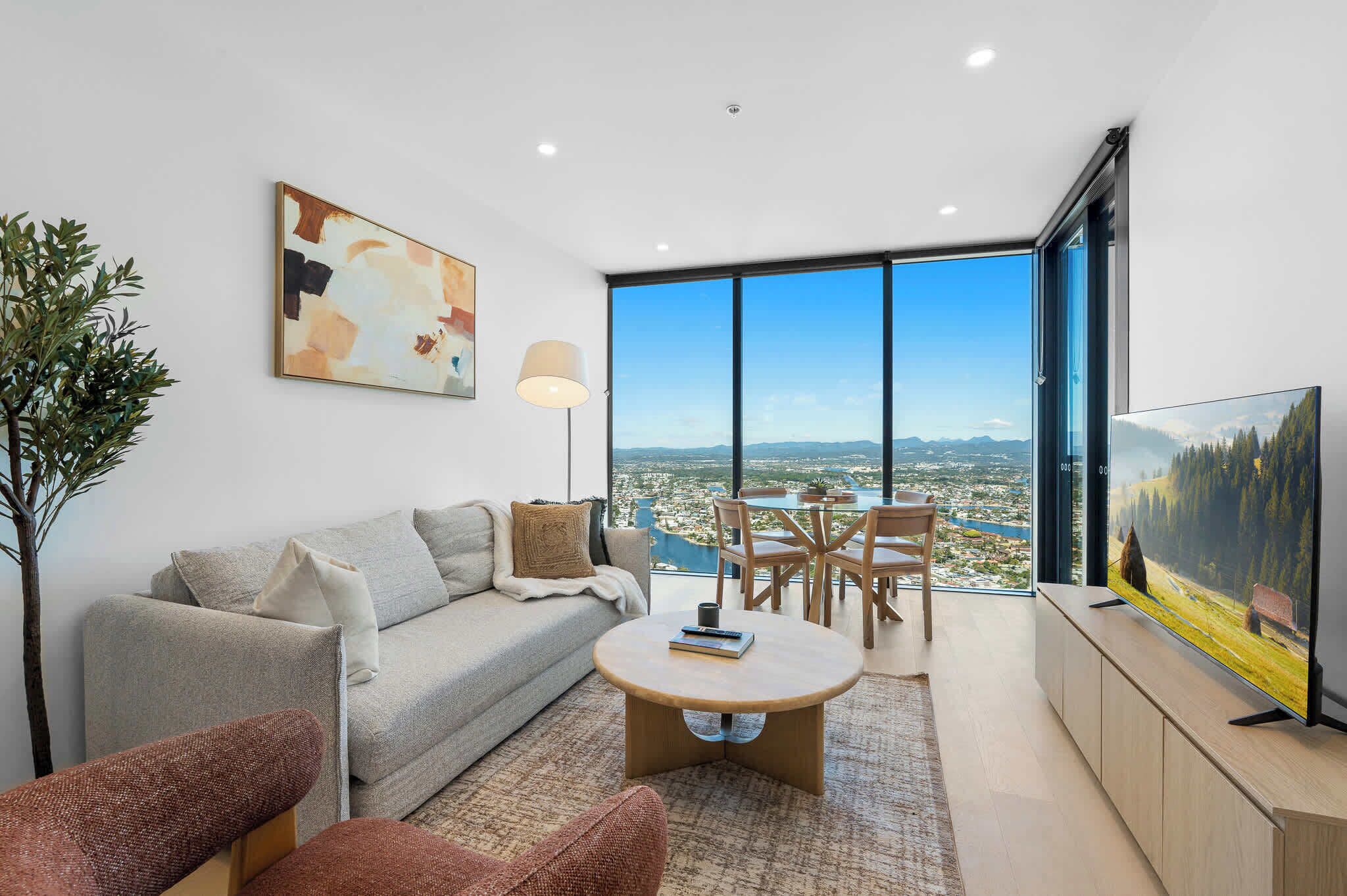 Un salon moderne avec un canapé, une table basse et des fauteuils, une grande fenêtre donnant sur la ville et un tableau accroché au mur.