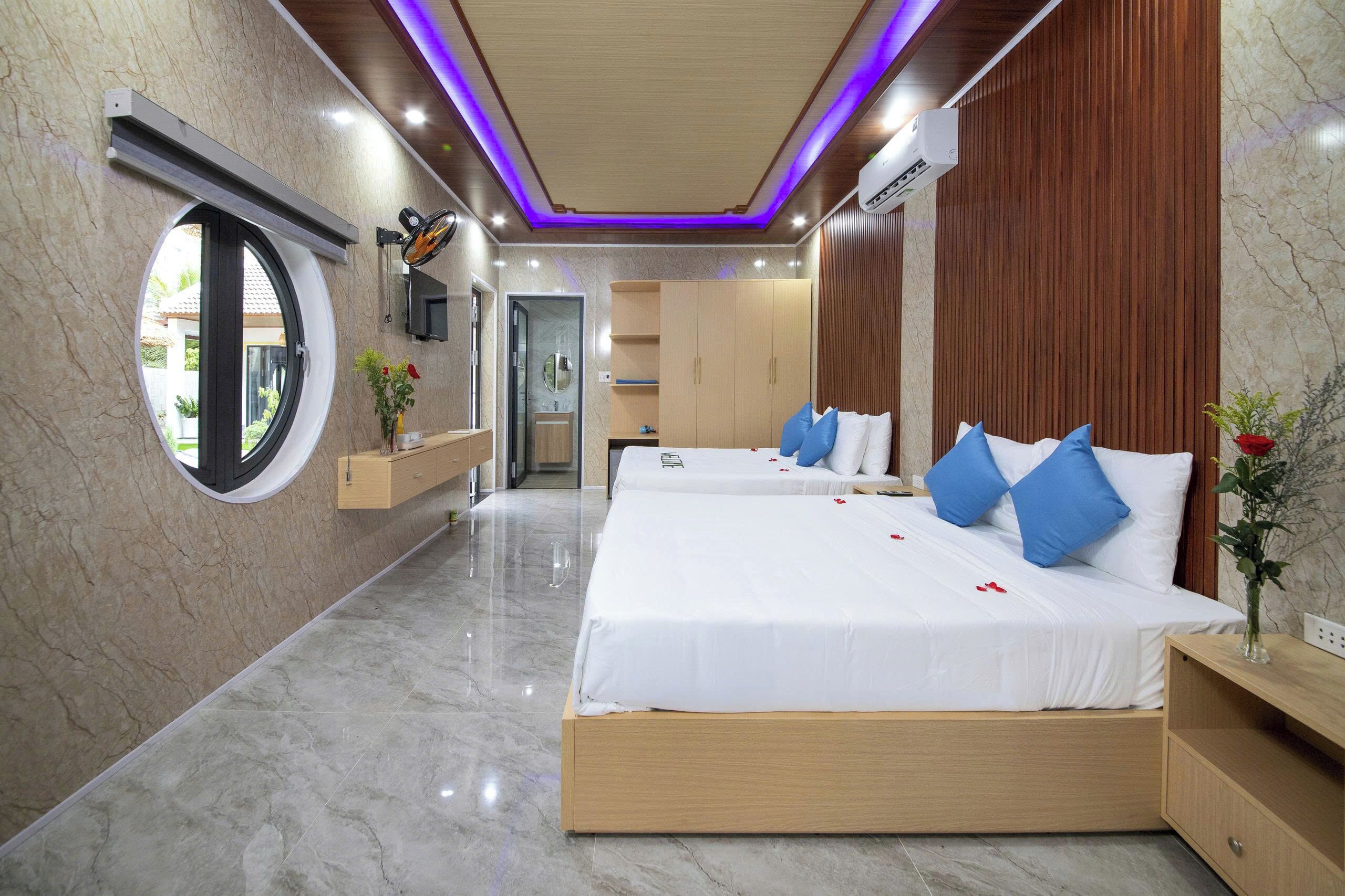 Habitación de hotel con dos camas, un ventanal redondo, un armario de madera y un baño con espejo.