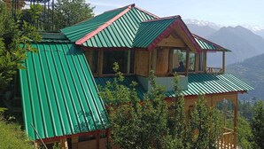 Una casa de dos pisos con techo de metal verde, estructura de madera y vista a las montañas al fondo.