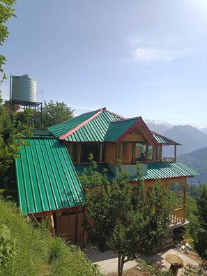 A two-story house with a green metal roof, wooden structure, and a view of mountains in the background.