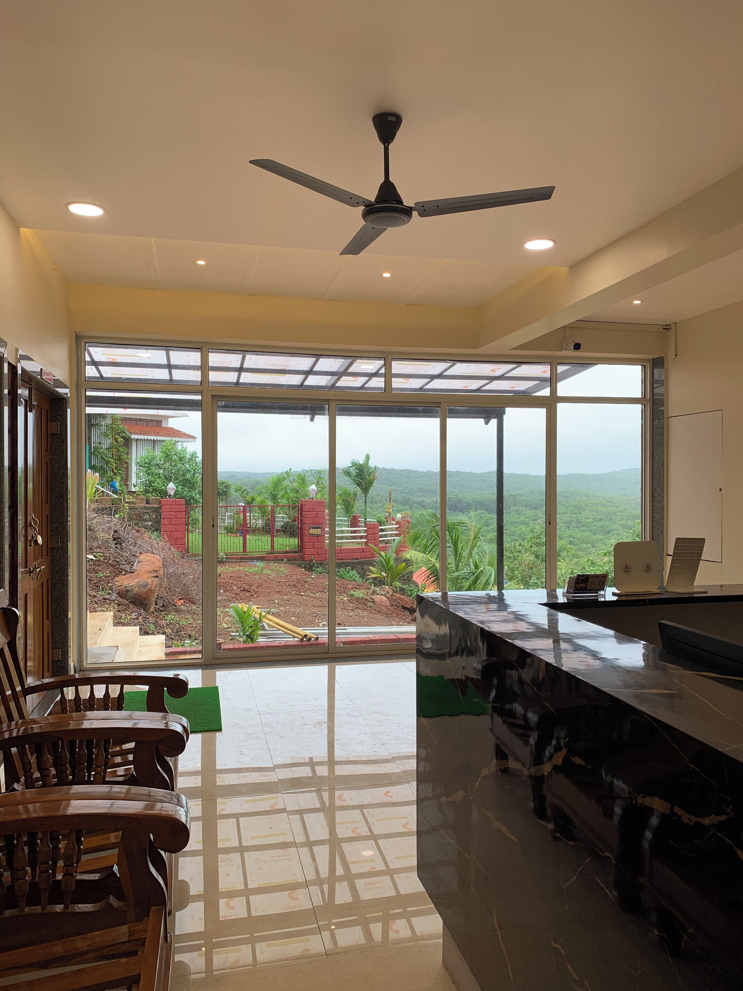 A spacious room with a ceiling fan, a large window offering a view of a lush landscape, and a bar area with a laptop and a magazine.