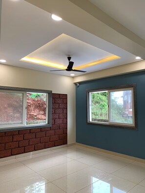 A room with a ceiling fan, two windows with views of greenery, a brick wall, and a tiled floor. - Anandi Hotel Guhagar (Guhagar)
