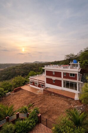 A two-story building with a flat roof, surrounded by greenery and hills. - Anandi Hotel Guhagar (Guhagar)