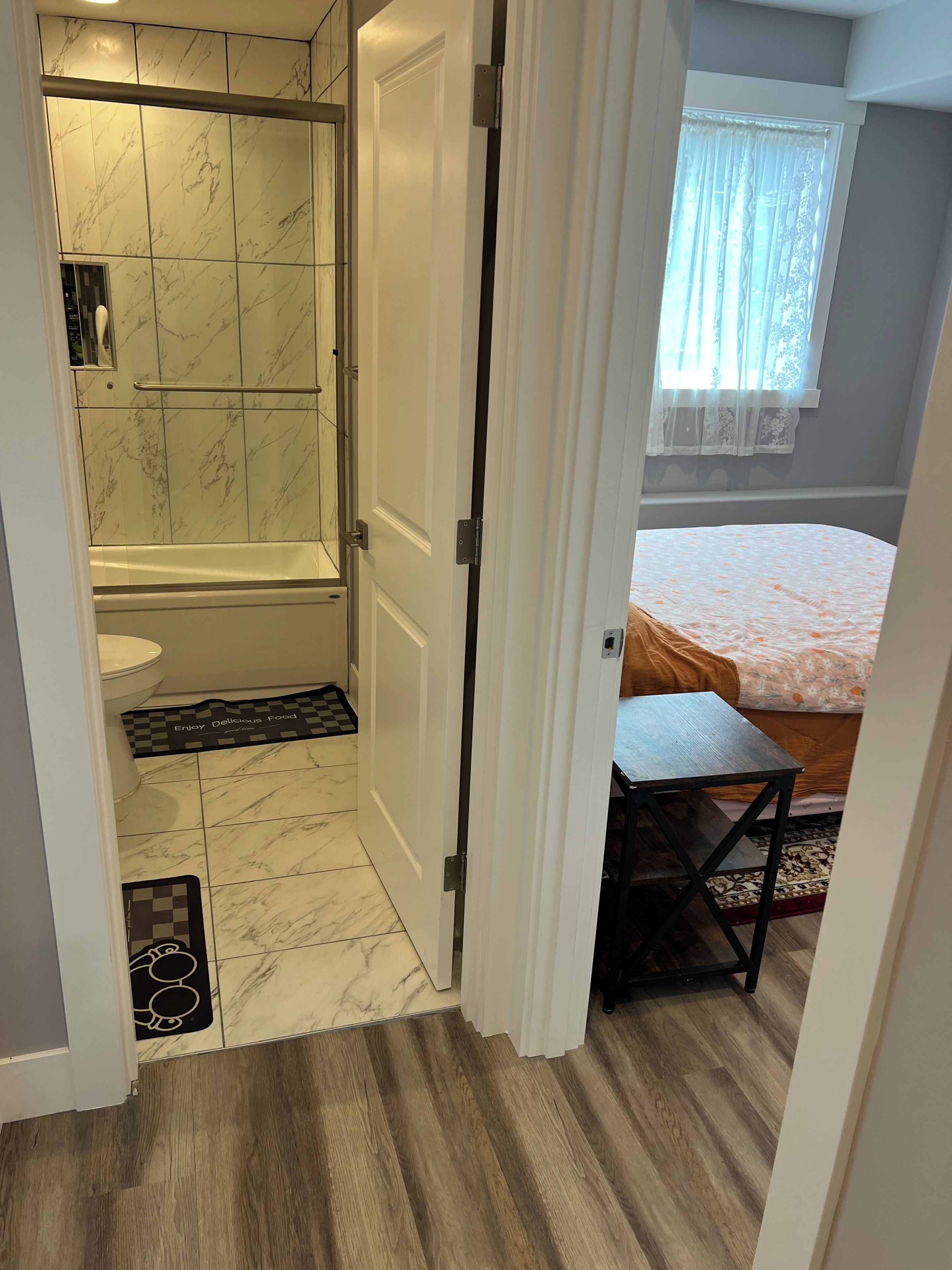 Ein Badezimmer mit Dusche, Toilette und Handtuchhalter. Im angrenzenden Zimmer befindet sich ein Bett mit einem gemusterten Überwurf.