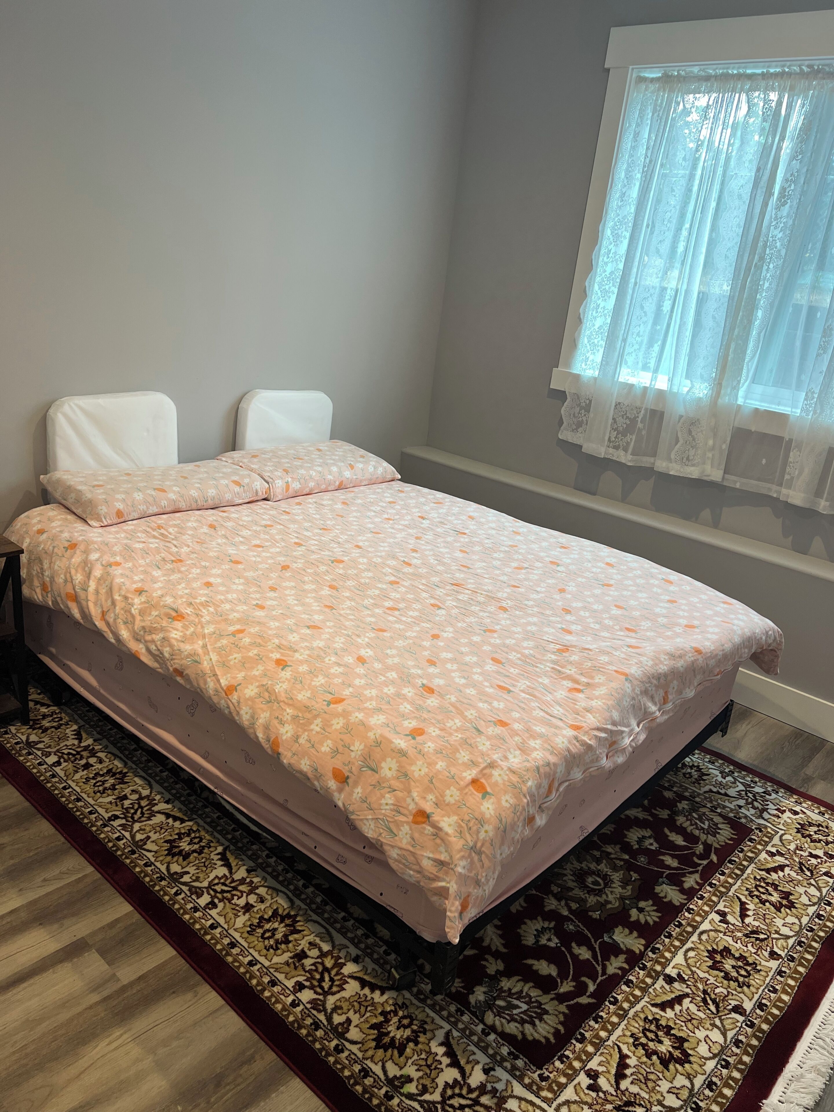 A bed with a floral bedspread, a patterned rug, and a window with sheer curtains.