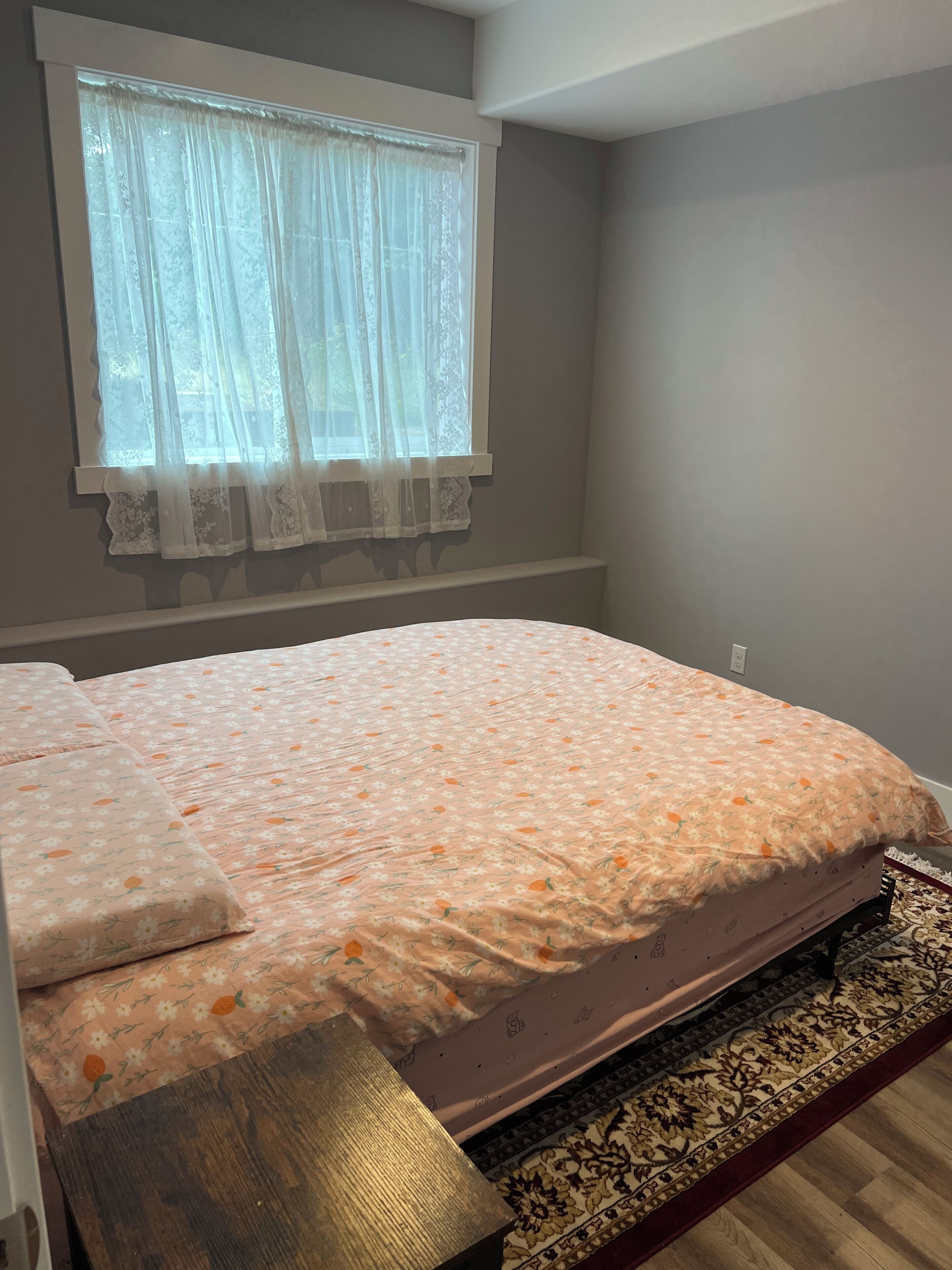 A bedroom with a bed, a window with sheer curtains, and a patterned rug.