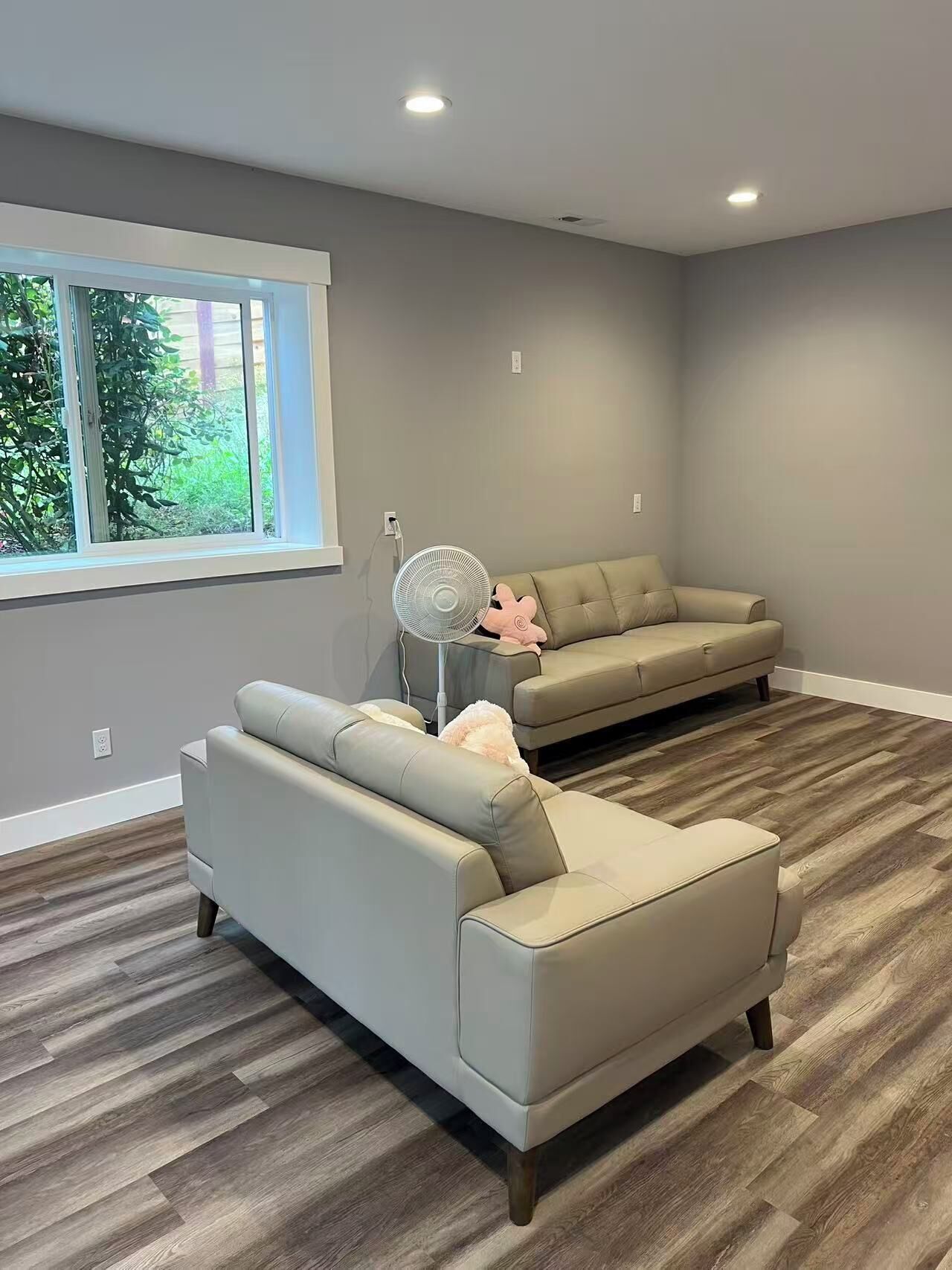 A modern living room with two sofas, a standing fan, and a large window.
