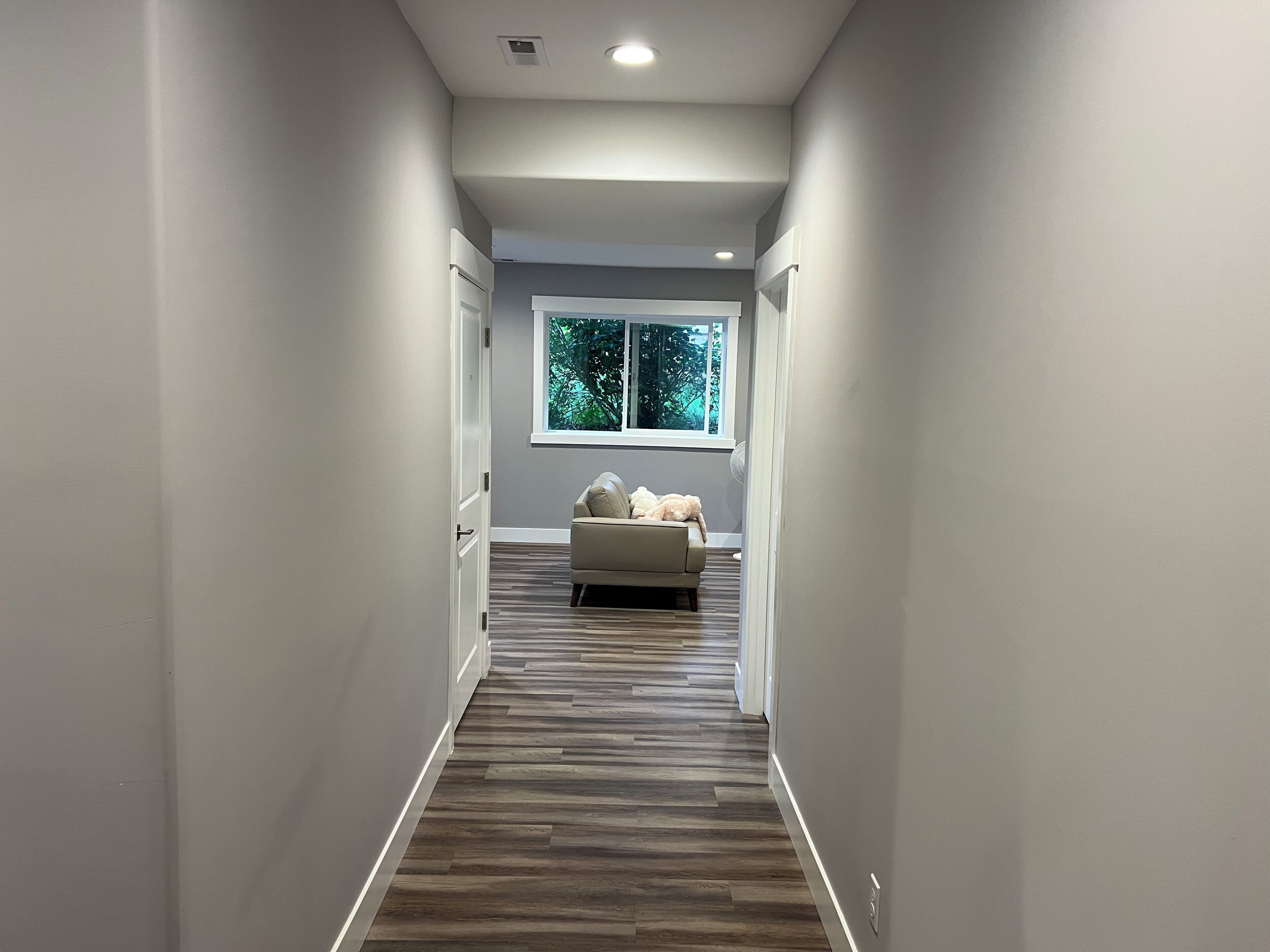 A hallway with a wooden floor, a window with a view of trees, and a white sofa with cushions.