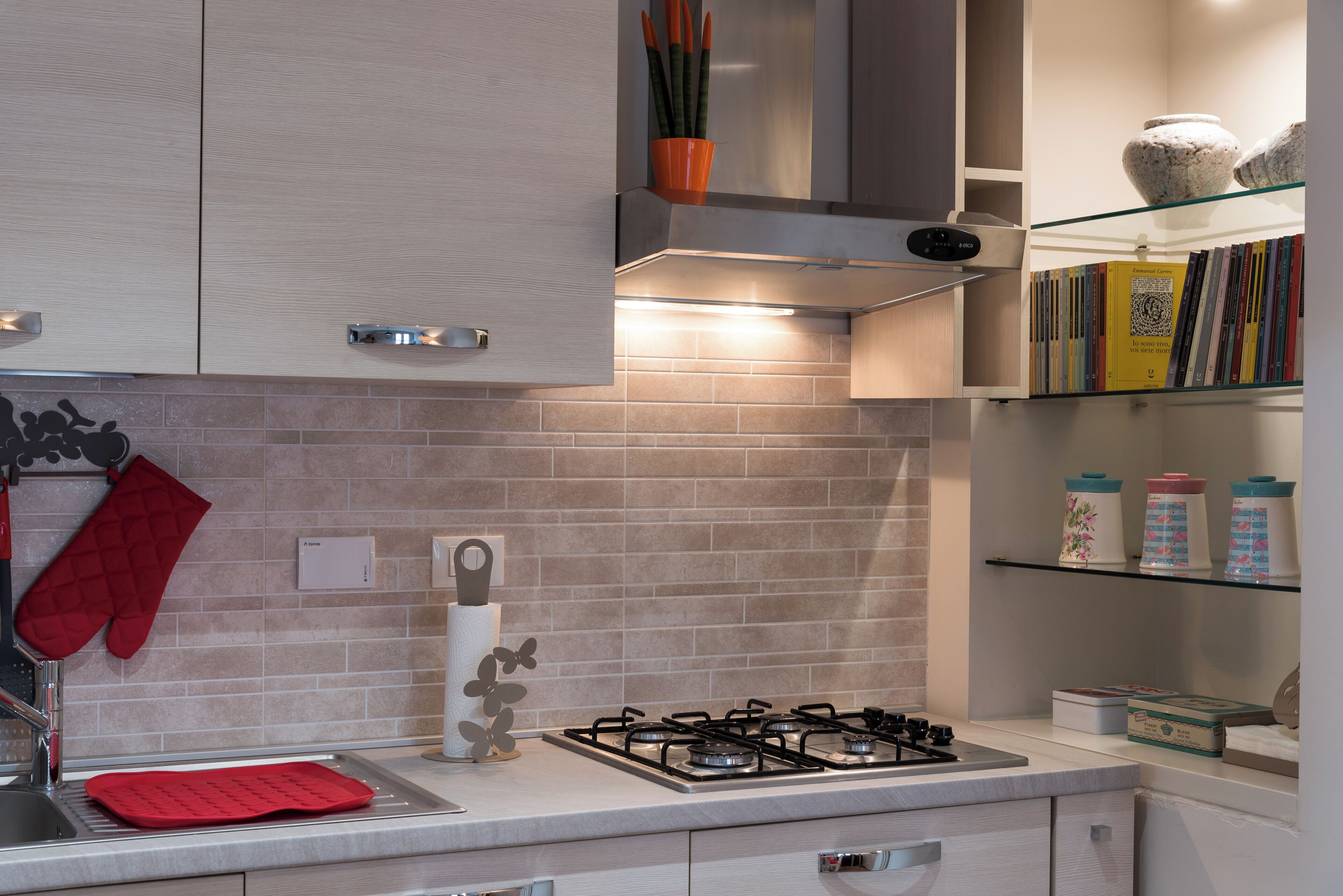 Una cucina moderna con piano cottura a gas, forno e cappa aspirante. Sono presenti armadietti, un piano di lavoro e uno scaffale con libri e utensili da cucina.