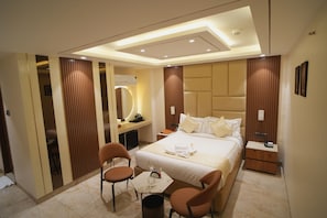 A modern hotel room with a large bed, a desk, two chairs, a vanity with a mirror, and a ceiling light.
