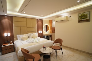 A hotel room with a large bed, bedside tables, a desk, two chairs, an air conditioning unit, and a painting on the wall.