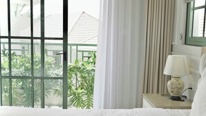 A hotel room with a bed, a nightstand with a lamp, a window with curtains, and a view of greenery outside. - Ngoc Khanh Villas Bai Khem Phu Quoc (Phu Quoc)